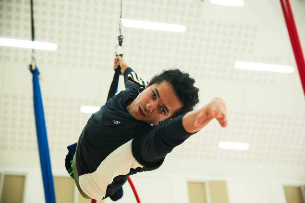 A photograph of Milton Lopes, a mixed race man, with shoulder length dreadlocks and who doesn't have fingers on his left hand. He is doing aerial harness, reaching towards the centre of the image with his left hand.