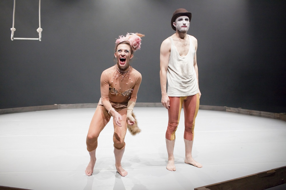 Two performers stand at the front of a round white stage. There is a trapeze hanging in the background. The performer on the left is laughing hysterically with their knees bent and is wearing a long sleeved unitard, with sequins and gold fabric sewn on to it, as well as a fur tail and a feather headdress. The performer on the right looks sad and is standing upright. They have clown-style face paint and are wearing a bowler hat, a white vest, and red and yellow leggings.