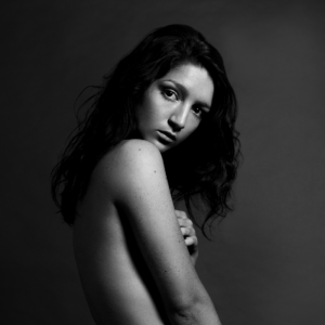 A black and white portrait image of the artist. She is slim, bare chested and wearing shorts with a bold white waistband. Her body is turned to the left, almost away and she is looking back directly at the camera with a neutral expression.
