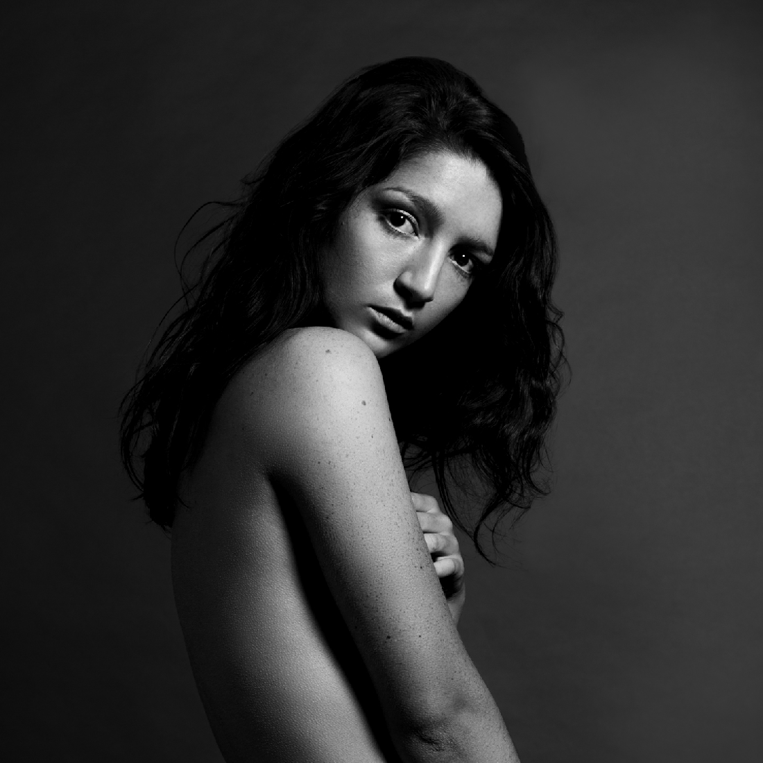 A black and white portrait image of the artist. She is slim, bare chested and wearing shorts with a bold white waistband. Her body is turned to the left, almost away and she is looking back directly at the camera with a neutral expression.
