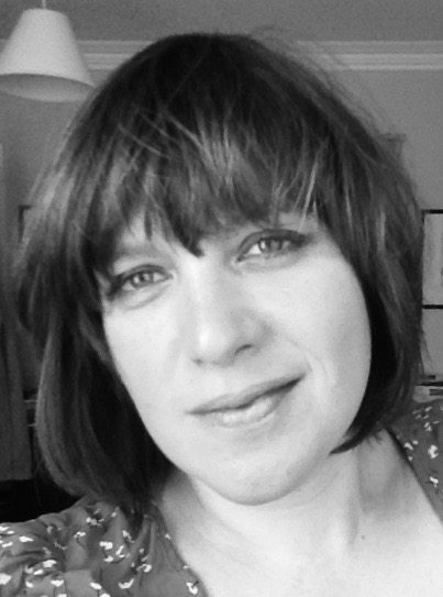 A head and shoulders balck and white portrait of Daisy Drury, who is a white woman with a blunt fringe and shoulder length dark hair. She is smiling and wearing a spotty blouse and subtle smokey eye make-up.