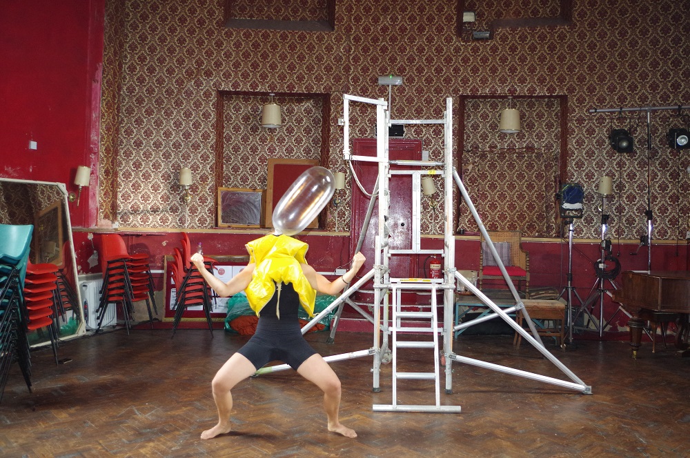 A photograph of Chloe during a residency. She is standing in the middle of a wallpapered room with a scaffolding tower in the background. She is in a wide stance, knees bent, arms wide forming bicep curls. A yellow life jacket is over her head and an inflated condom is emerging in a long balloon from the place where her head should be.