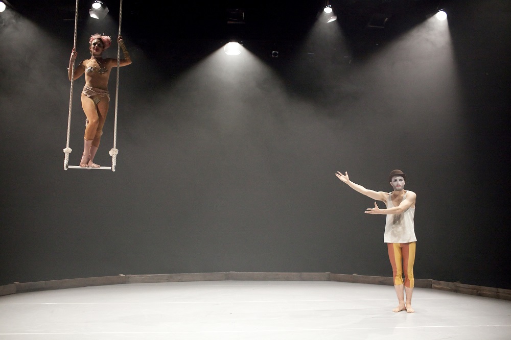 There is a round white stage filled with hazy smoke. On the lft there is a performer wearing a long sleeved unitard, with sequins and gold fabric sewn on to it, as well as a fur tail and a feather headdress, who is standing on a trapeze rigged from the ceiling. On the right, there is a performer with clown-style face paint and are wearing a bowler hat, a white vest, and red and yellow leggings, who is gesturing towards the performer on the trapeze, as if to say ‘Ta-da’.