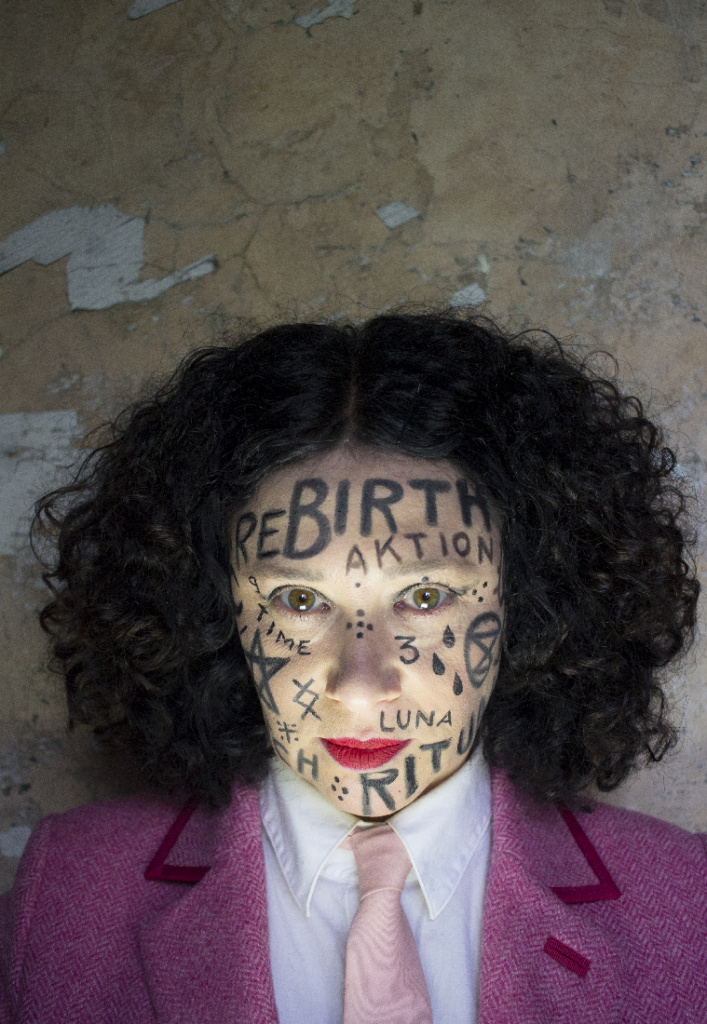 A photograph of a woman with curly brown shoulder length hair wearing a pink tweed suit jacket, white shirt and pink tie. The words rebirth, aktion, luna, time and ritual are written on her face with black crayon, along with various drawings and symbols.