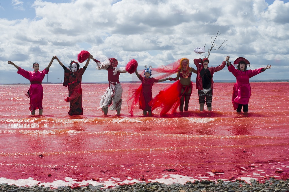 Seven people stand in the shallows of a red sea in front of a blue and cloudy sky. The people are wearing different red costumes and are holding hands together in the air.