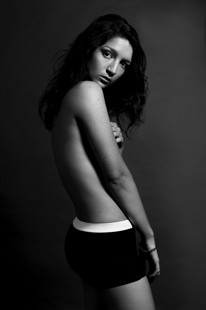 A black and white portrait image of the artist. She is slim, bare chested and wearing shorts with a bold white waistband. Her body is turned to the left, almost away and she is looking back directly at the camera with a neutral expression.