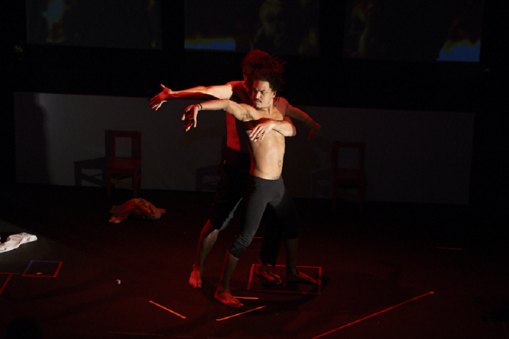 Photo of live circus-theatre production What The Circus? A white man in his 40’s and a younger, slightly shorter, black man dance together in the middle of a black stage under a red stage light. Standing close, the taller man is behind, with his left arm tenderly placed onto the smaller man's chest. They are both looking to the right with their right arms extended in the same direction, their palms are open, thrusting forward, fingers stretching toward the floor. White tape marks out squares and lines under their feet, and two items of clothing are strewn on the floor beside them.