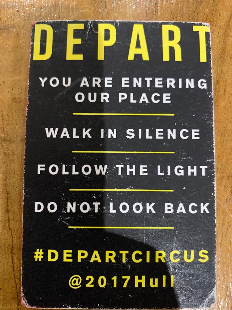 A photograph of a card distributed to audiences at the Depart shows. The paper is slightly worn. Yellow writing in block letters on a black background state You are entering our place. Walk in silence. Follow the light. Don’t look back. #departcircus @2017Hull