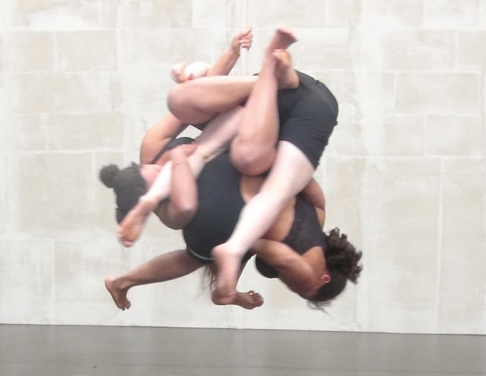 Three human bodies, with white and brown skin, are seemingly merged together, to become a “ball” of different heads and limbs suspended approximately two feet above the ground. You can not see the faces on the individual performers, they appear to exist as one entity. The wall behind is made of sand coloured bricks and the floor is grey, and has a large crack pictured in the foreground of the image.